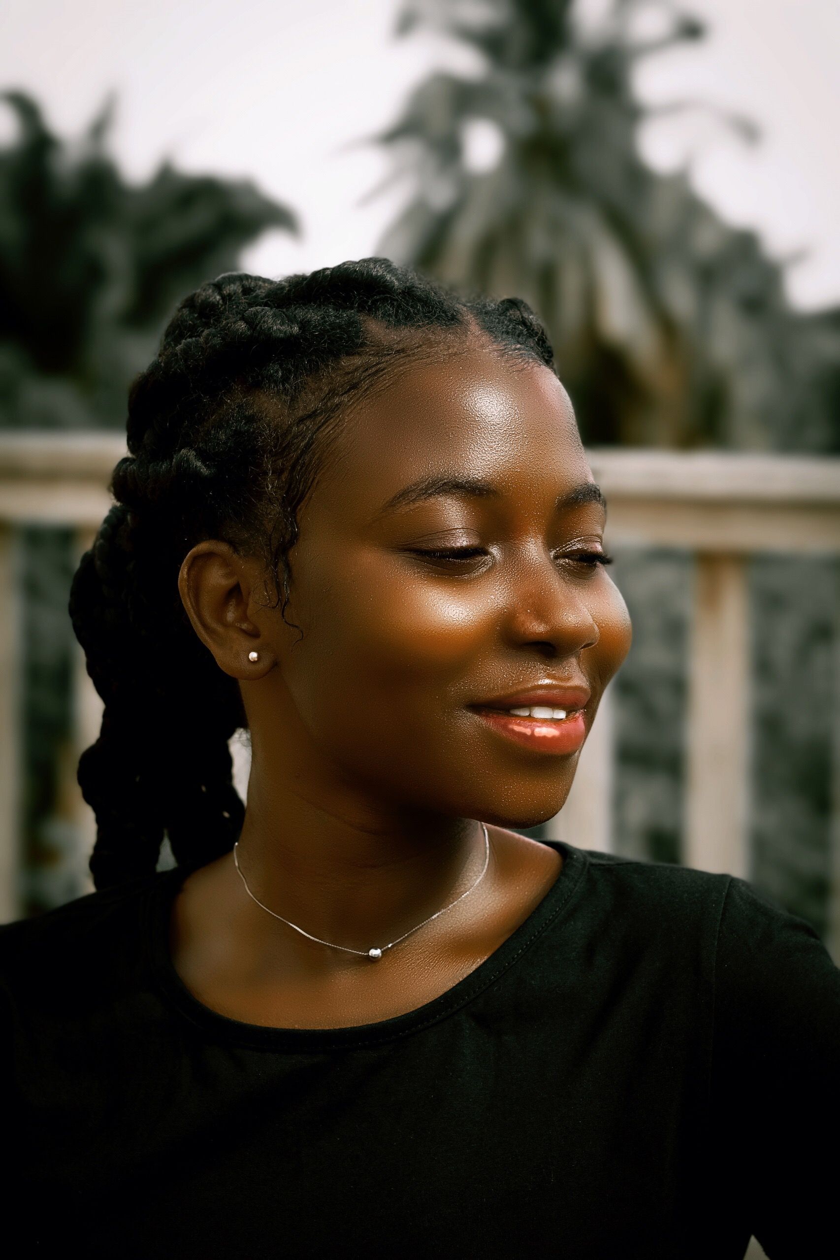 Striking portrait of a young African woman with braided hair and a necklace, smiling outdoors with blurred trees in the background.